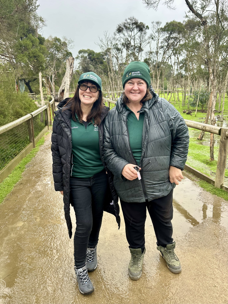 Two women, both smiling at the camera and wearing green uniforms with beanies and jackets stand on a wet pathway with trees in the background.